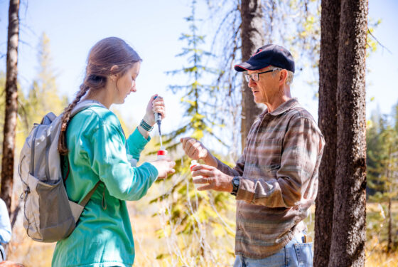A professor demonstrates how to inject a water sample to a student as they stand outside in front of a pond.