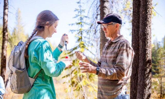 A professor demonstrates how to inject a water sample to a student as they stand outside in front of a pond.