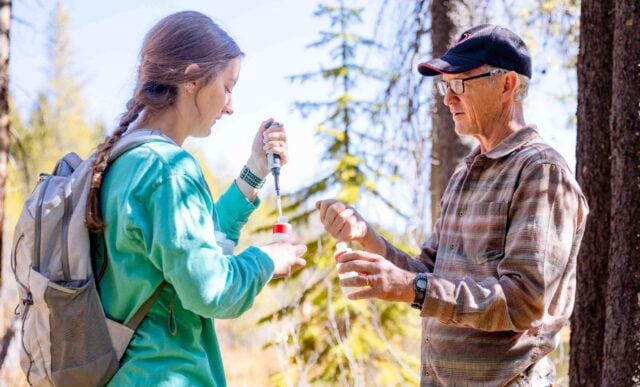 A professor demonstrates how to inject a water sample to a student as they stand outside in front of a pond.