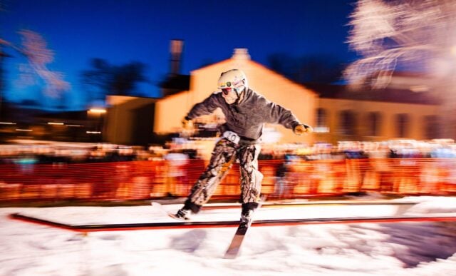 A skier slides a rail in front of the library while students cheer for him in the background. A motion blur effect is used in this photo.