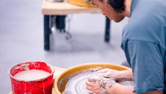A student is seated at a potter's wheel and starts forming a pot with clay.