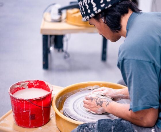 A student is seated at a potter's wheel and starts forming a pot with clay.