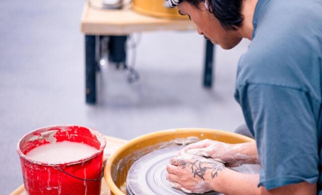 A student is seated at a potter's wheel and starts forming a pot with clay.