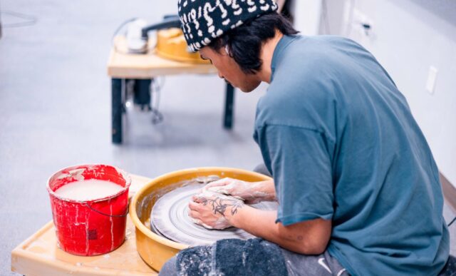 A person wearing a patterned beanie and a blue shirt is shaping clay on a pottery wheel while seated at a workbench with a red bucket of water nearby.