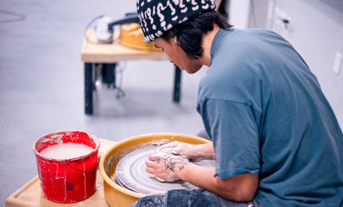A student is seated at a potter's wheel and starts forming a pot with clay.