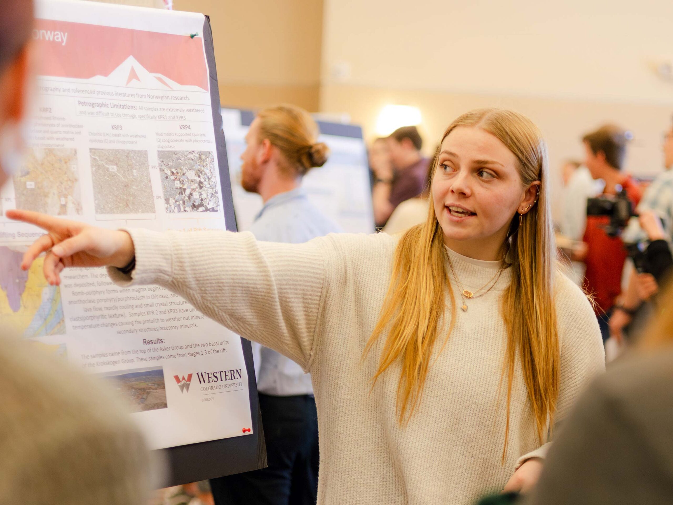 A student points at her research presentation that is displayed on a large poster board.