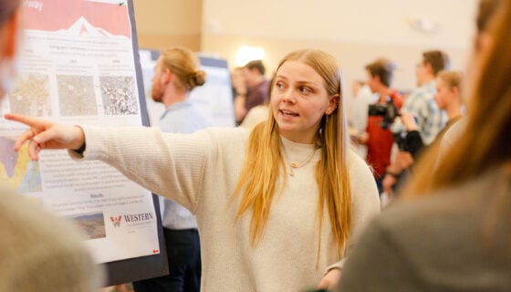 A student points at her research presentation that is displayed on a large poster board.