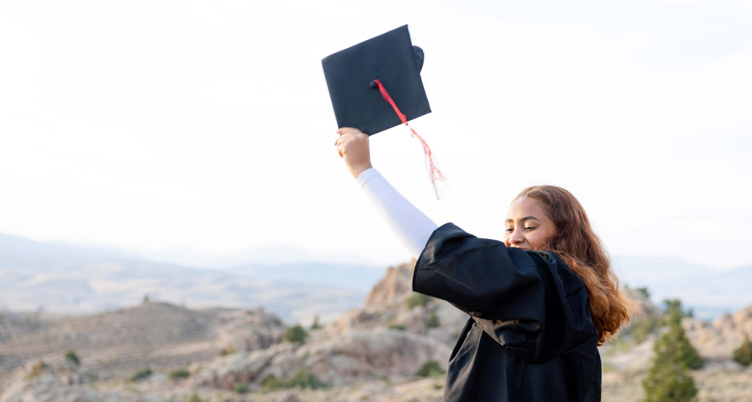 A student wears a graduation gown and holds her cap in the air at Hartman Rocks.