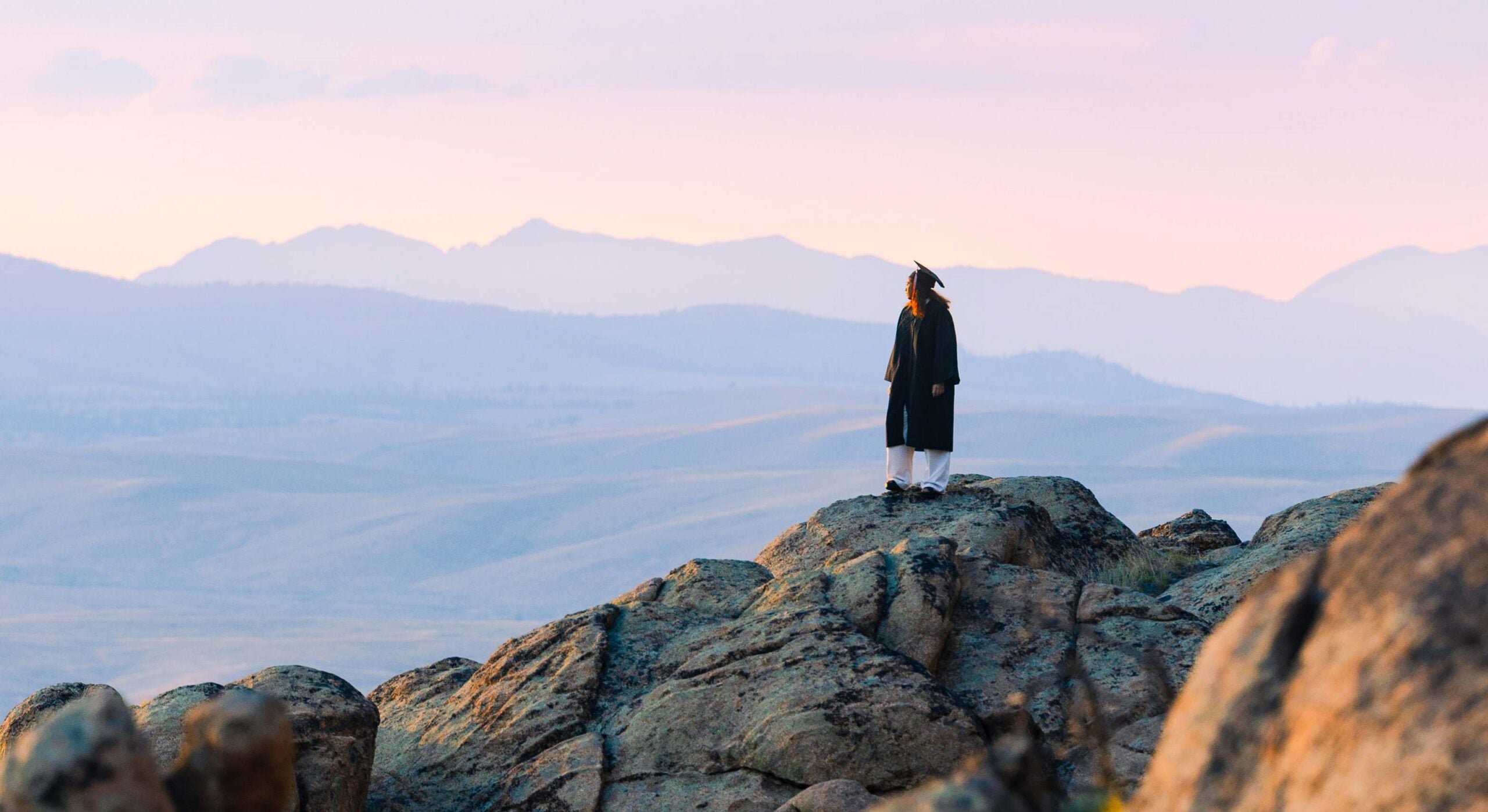 A student wearing a cap and gown stands on a Hartman Rock formation at sunset.