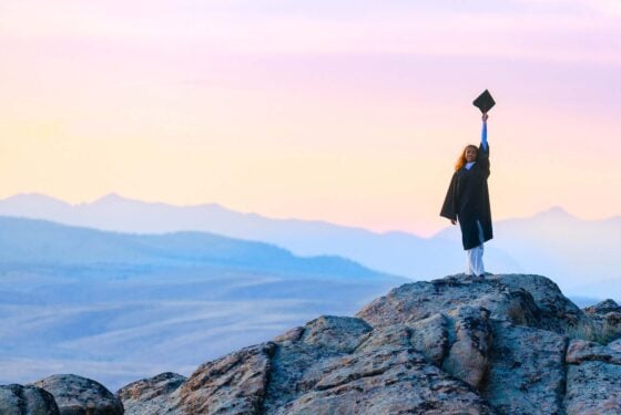 A student wearing a graduation gown stands on a rock feature at Hartman Rocks and holds her cap up at sunset with big mountain views behind her.