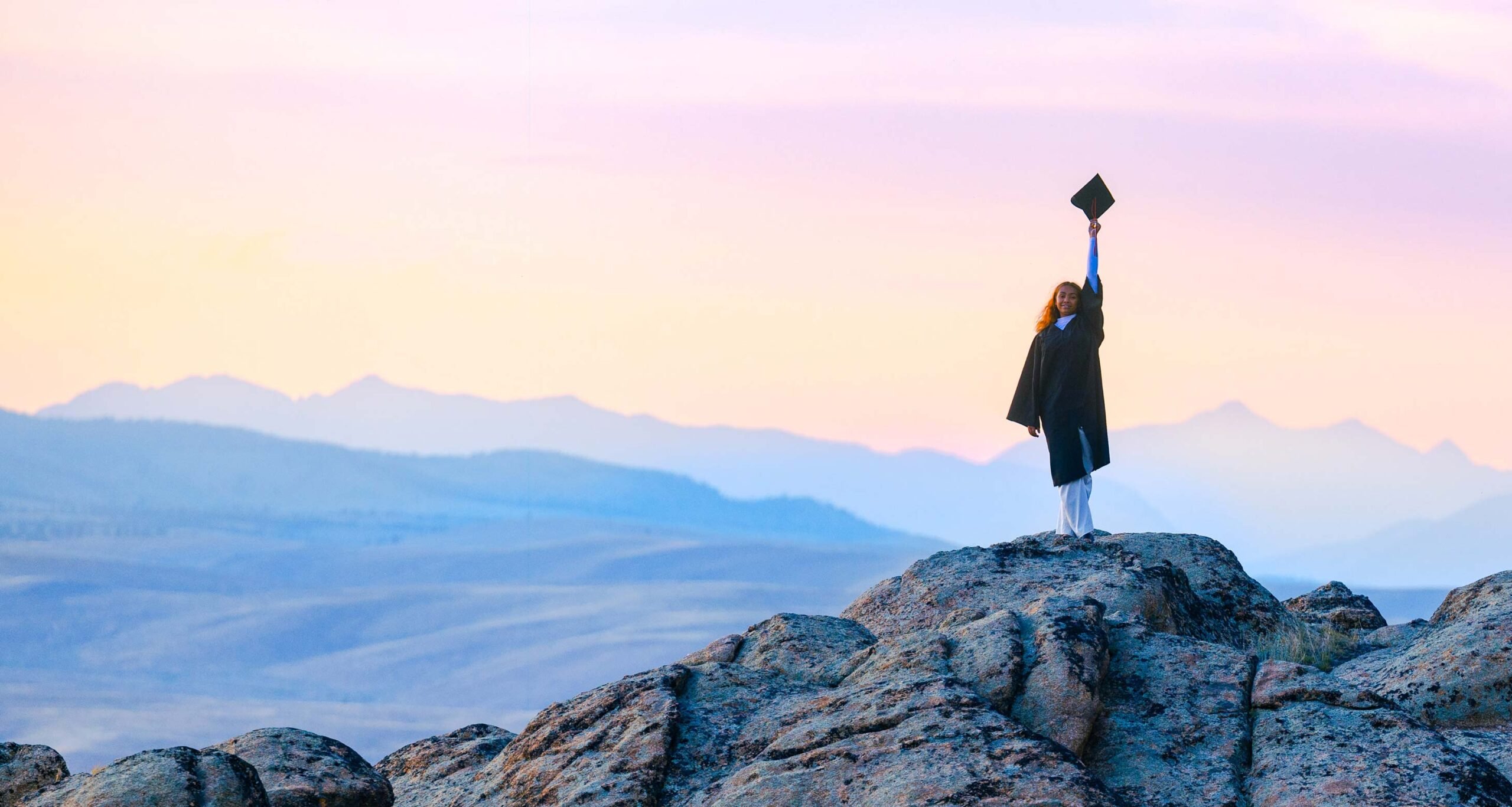 A student wearing a graduation gown stands on a rock feature at Hartman Rocks and holds her cap up at sunset with big mountain views behind her.
