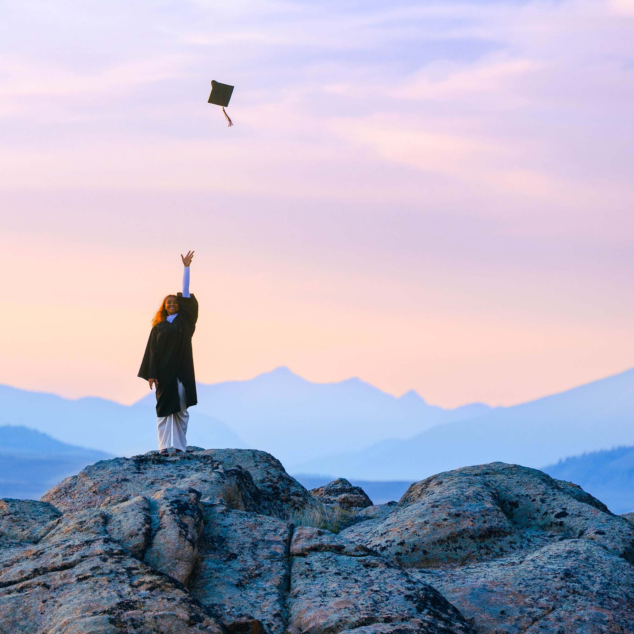 A student wearing a graduation gown stands on a rock feature at Hartman Rocks and throws her cap in the air at sunset with big mountain views behind her.