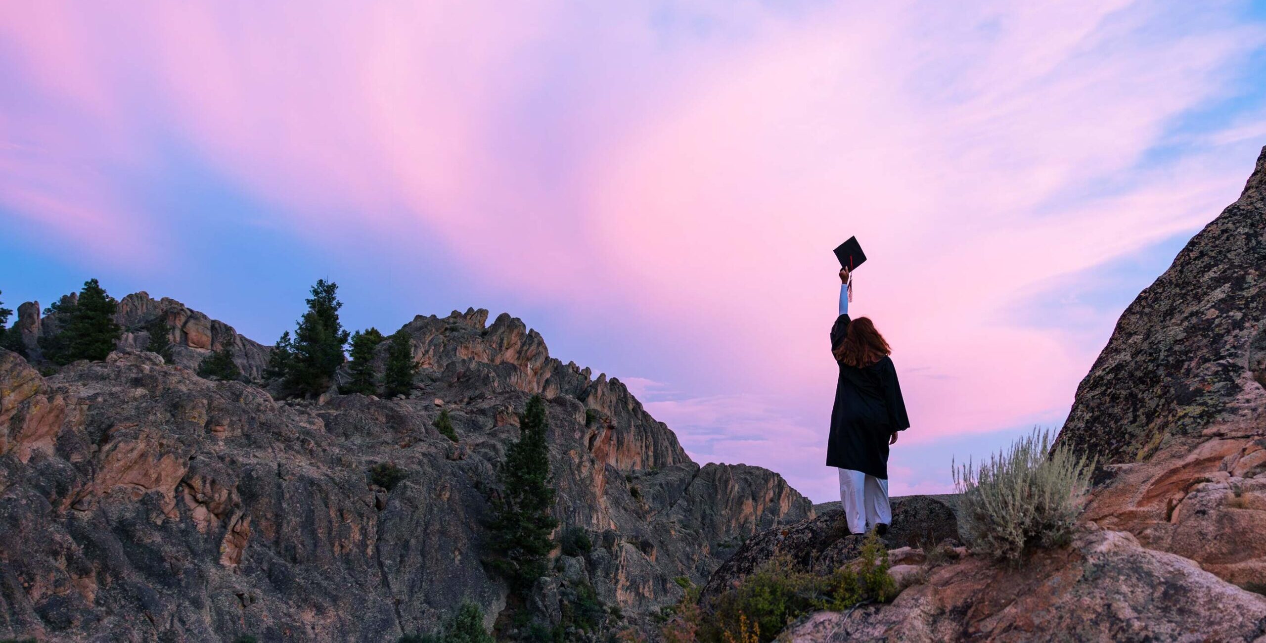 A girl wearing a graduation cap and gown raises her cap during a vibrant sunset at Hartman Rocks.