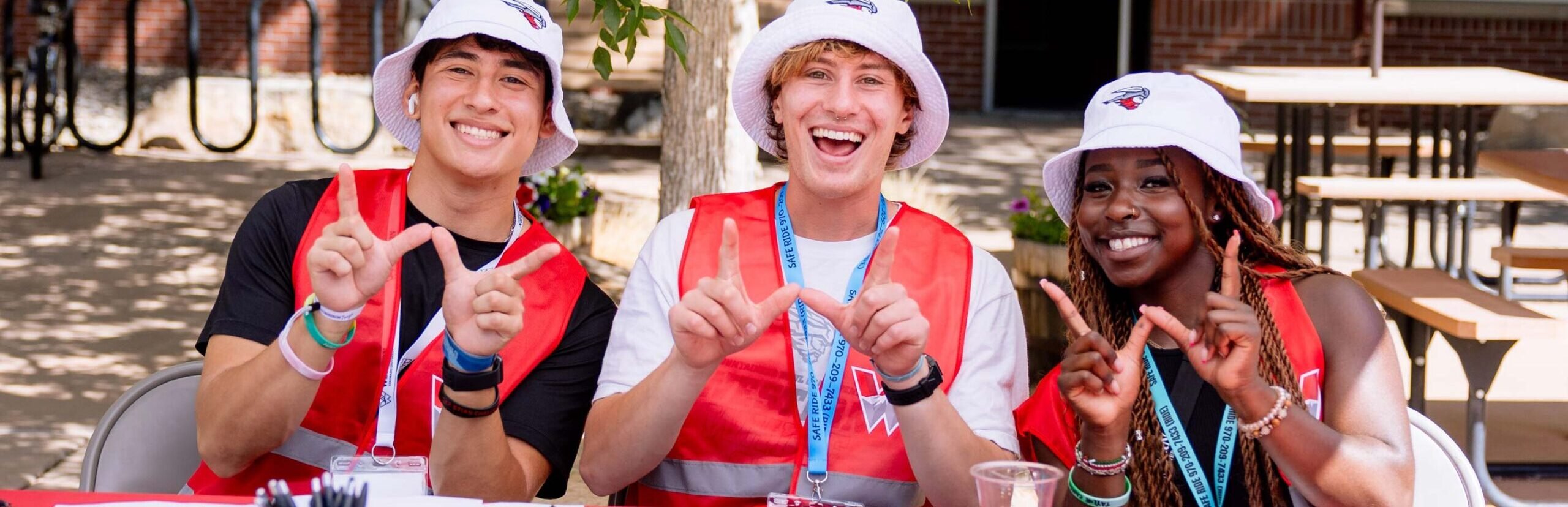 3 orientation leaders are seated at a check-in table and hold their hands up in a