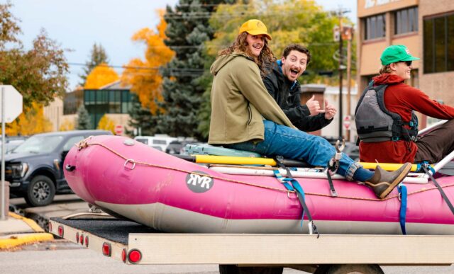 Three students sit on a trailer with a raft on it as they parade down Main Street during the Homecoming parade.