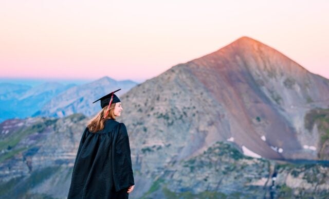 A student, looking off into the distance, wears a cap and gown as she stands on a high alpine ridge with a large peak behind her at sunrise.
