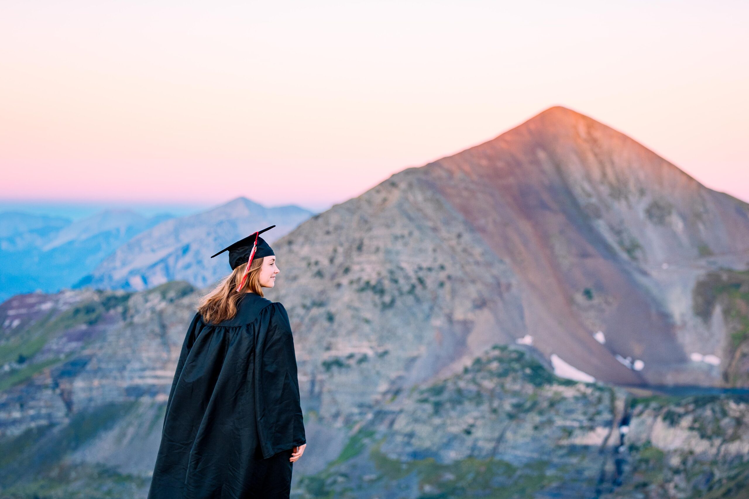 A student, looking off into the distance, wears a cap and gown as she stands on a high alpine ridge with a large peak behind her at sunrise.