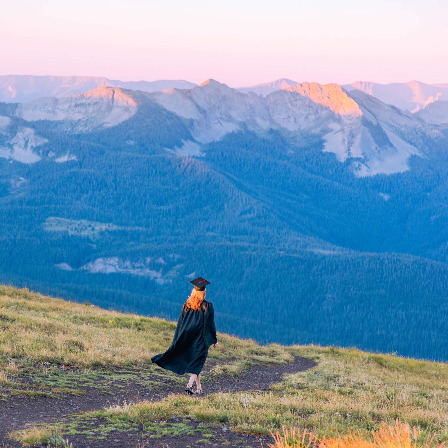 A student wearing a cap and gown walks down a high alpine ridgeline at sunrise.