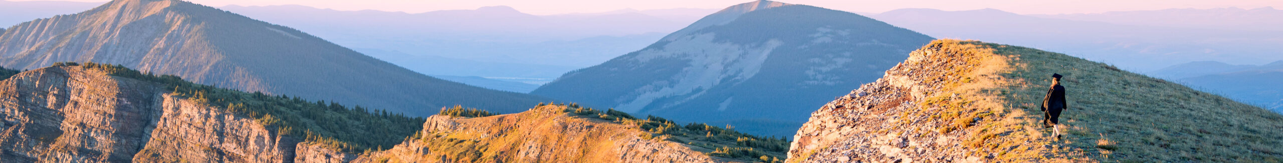 A student wearing a cap and gown walks down a high alpine ridgeline at sunrise. The ridge is lit by morning alpenglow.