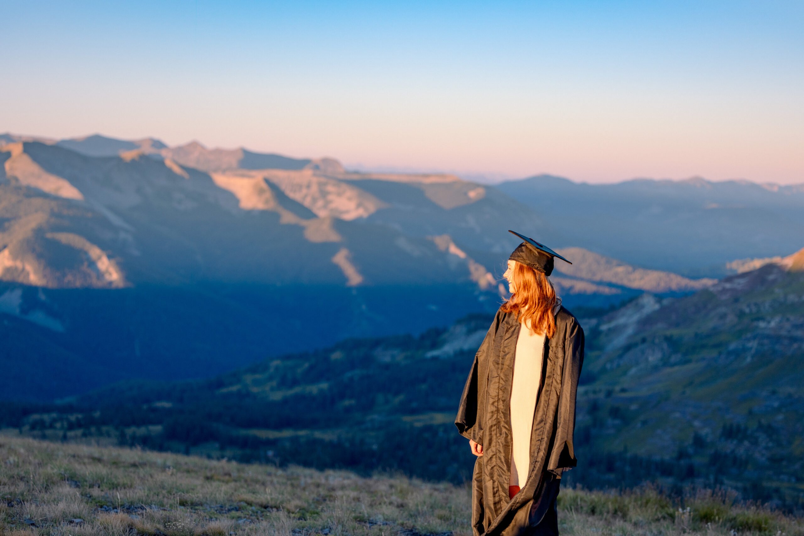 A student wearing a cap and gown smiles as she stands on a high alpine ridge at sunrise with big mountain views in the background.