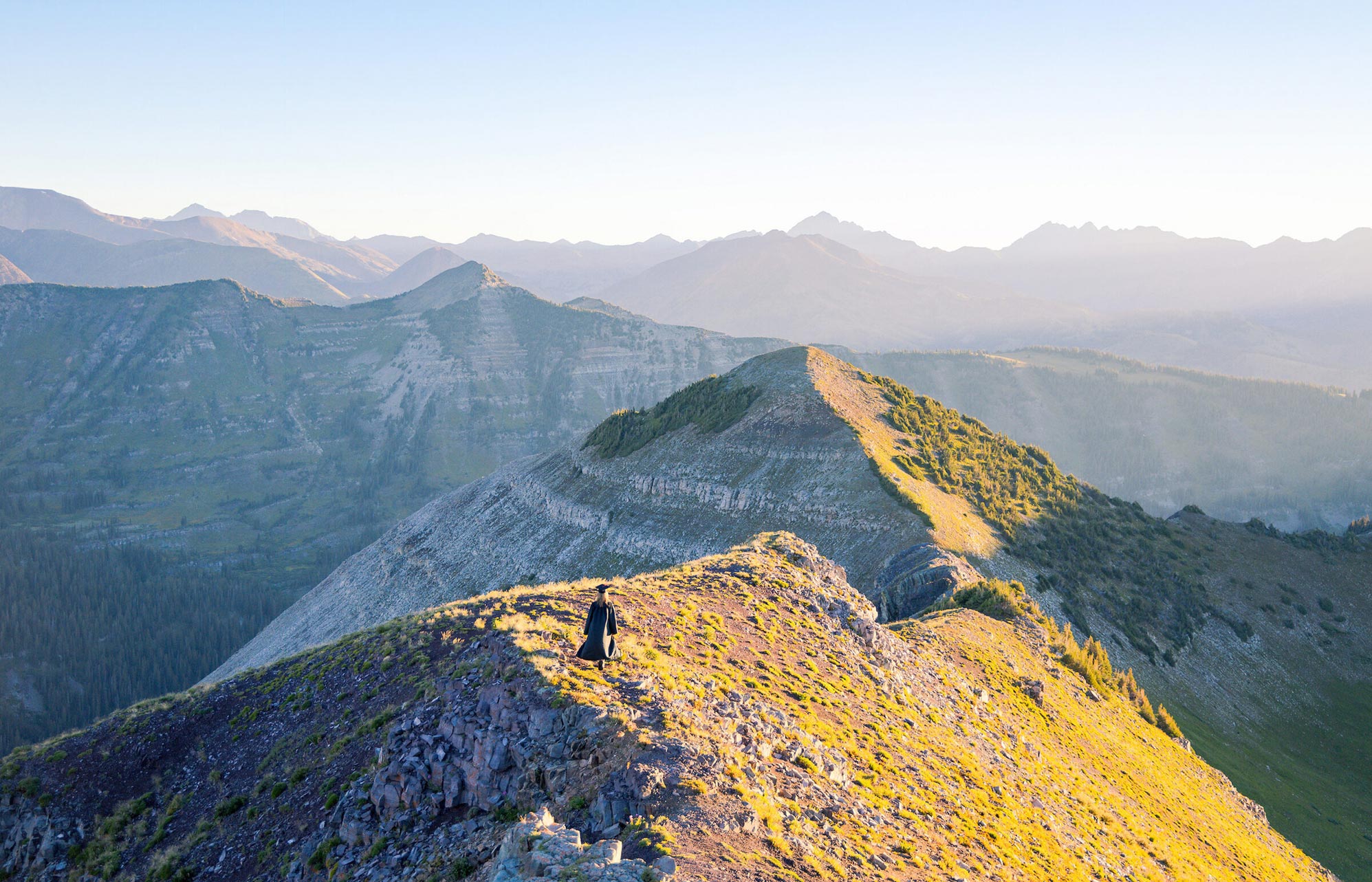 A student wearing a cap and gown walks down a high alpine ridge line at sunrise. The ridge is lit by morning alpenglow.