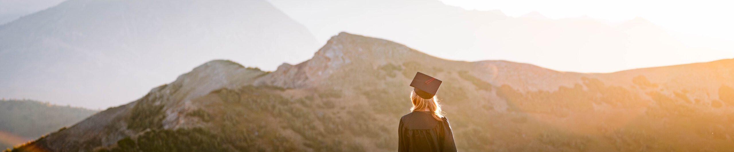 A student wearing a gown and raising her cap stands facing away from the camera on a high alpine ridge at sunrise with big mountain views glowing in the background.