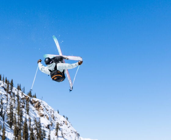 A skier cork 7s in the air on a blue sky day with mountains in the background.
