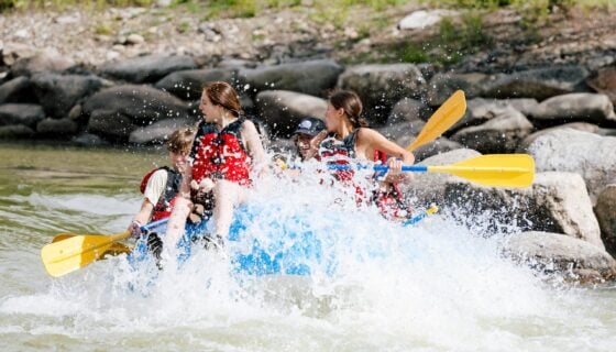 Students on a raft at the Whitewater Park are splashed by a rapid.