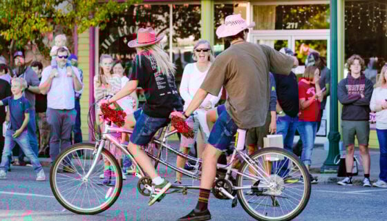 Two students wearing pink cowboy hats ride a tandem townie bike down main street and bring joy to community members during the Homecoming parade.