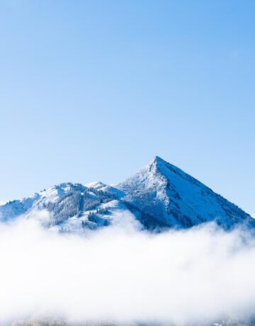 Mt. Crested Butte peeks through a low layer of clouds on a blue sky day.