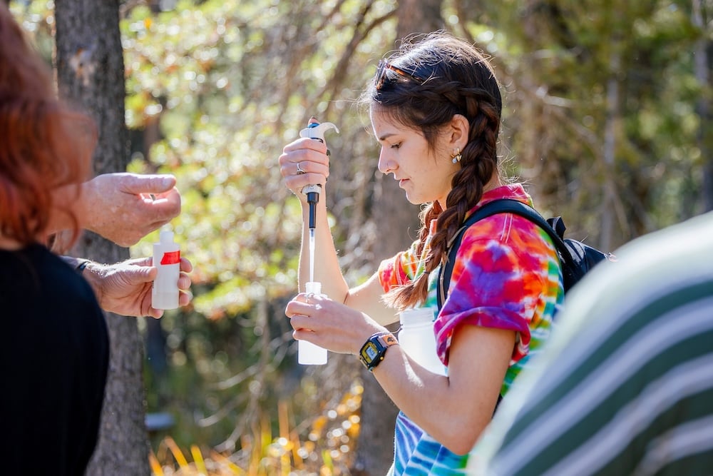 A student injects acid into a water sample as she stands in the forest wearing a tie dye shirt.