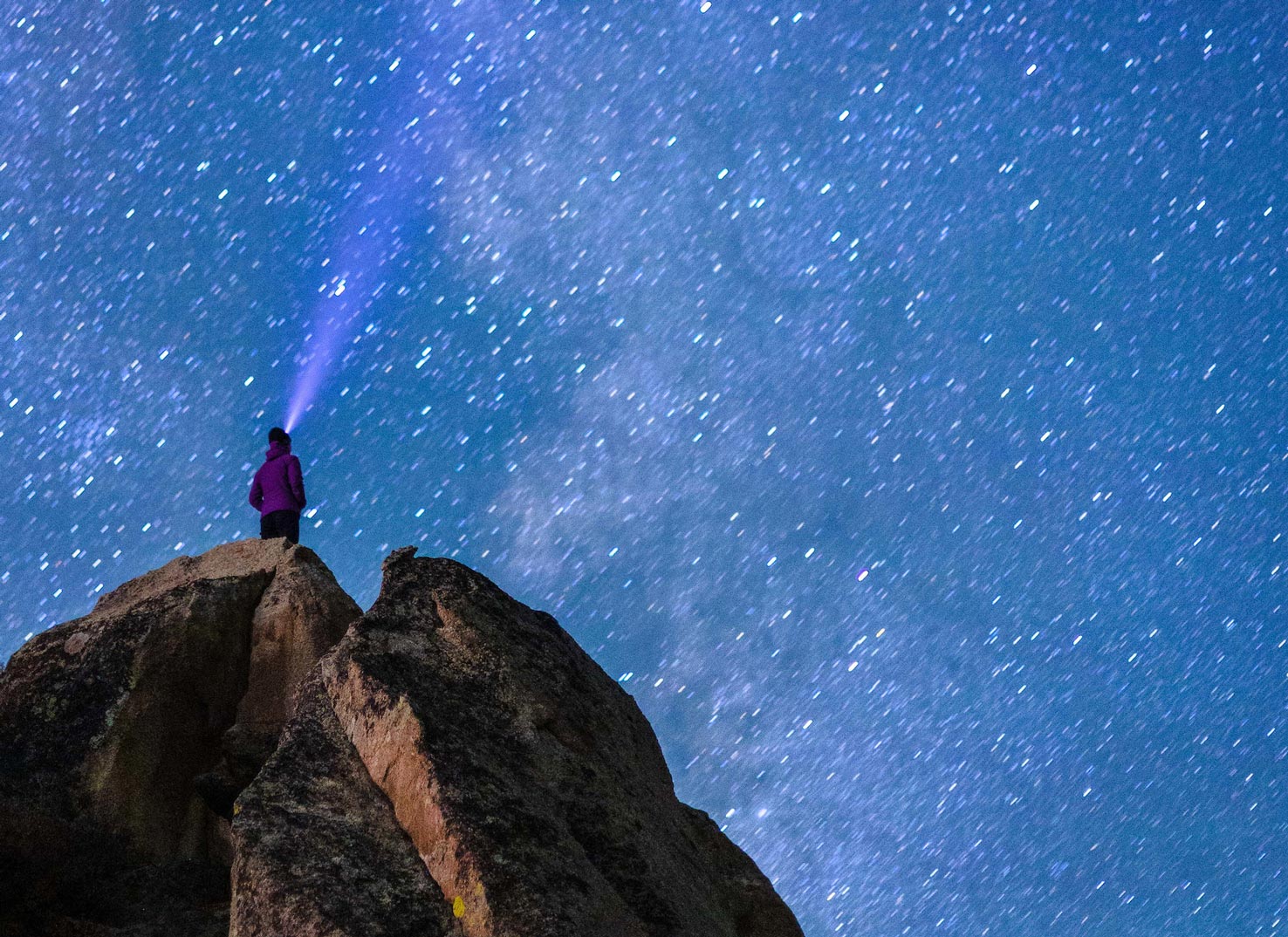 A girl with a head lamp stands on a rock and looks at the milkyway