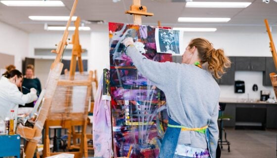 A student paints an abstract piece on a canvas inside a classroom.