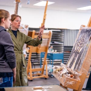 A professor gives feedback to her student next to her on her painting
