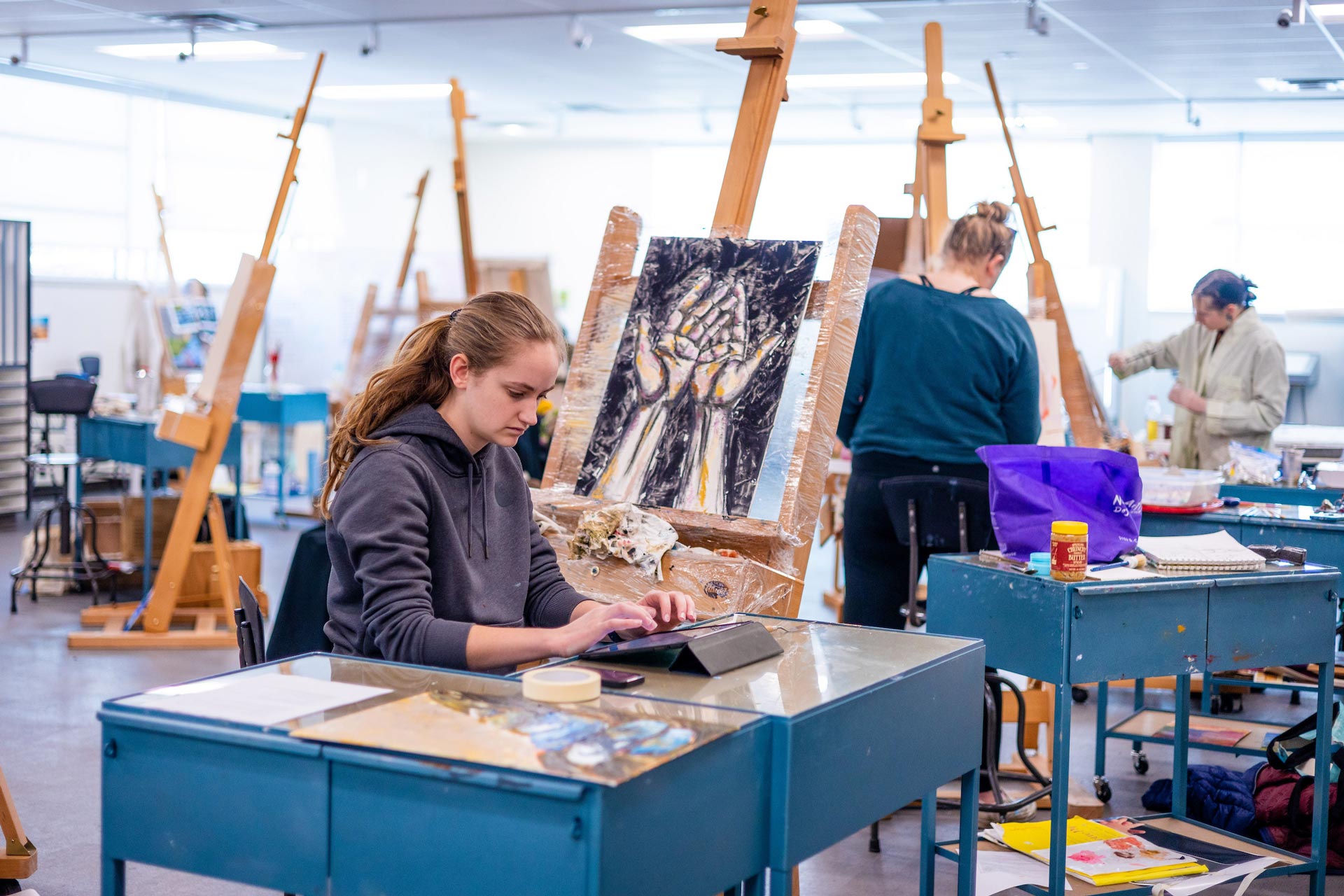 A student types on her tablet while seated in front of her painting of hands