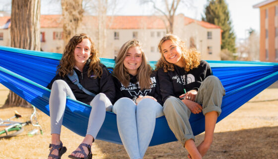 Three students sit on a hammock in front of a dorm complex.