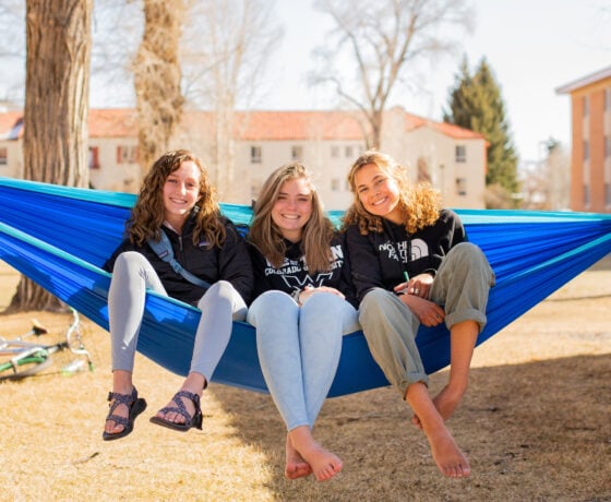 Three students sit on a hammock in front of a dorm complex.