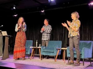 CMarie Furhman, Derek Sheffield, and Elizebeth Bradfield applaud while standing on a stage.