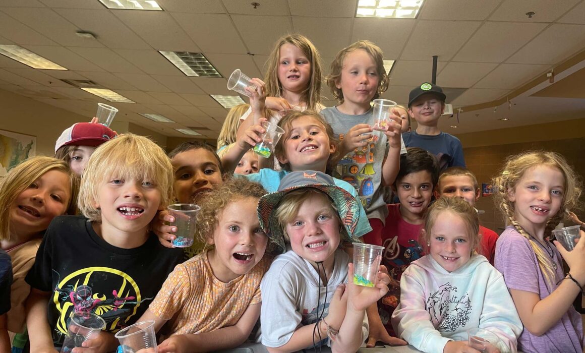 Children attending a Junior Mountaineer summer camp holding beakers inside a classroom.