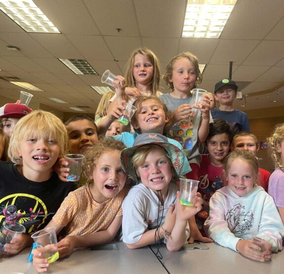 Children attending a Junior Mountaineer summer camp holding beakers inside a classroom.