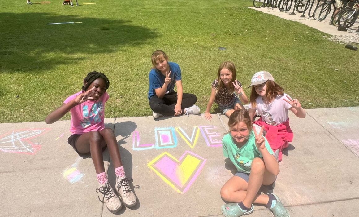 Five kids smile while creating chalk art on the campus sidewalks.
