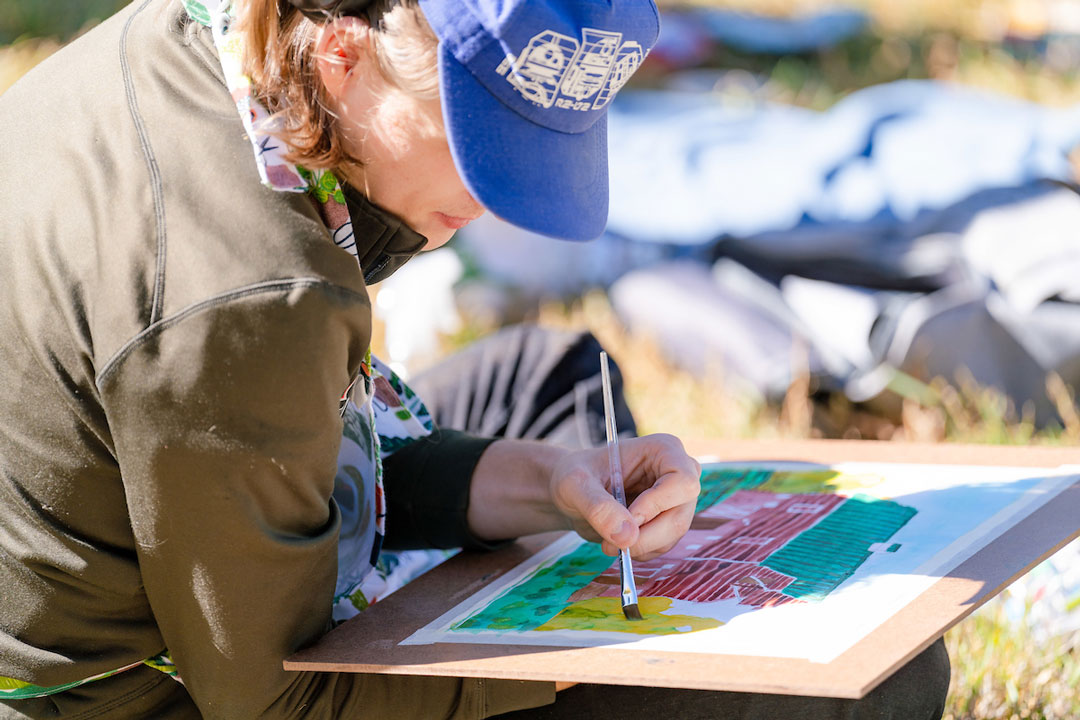 A close up of a student sittin gon grass and painting a barn with watercolors.