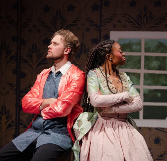 An actor and actresses sit on a stool with their backs turned away from each other.