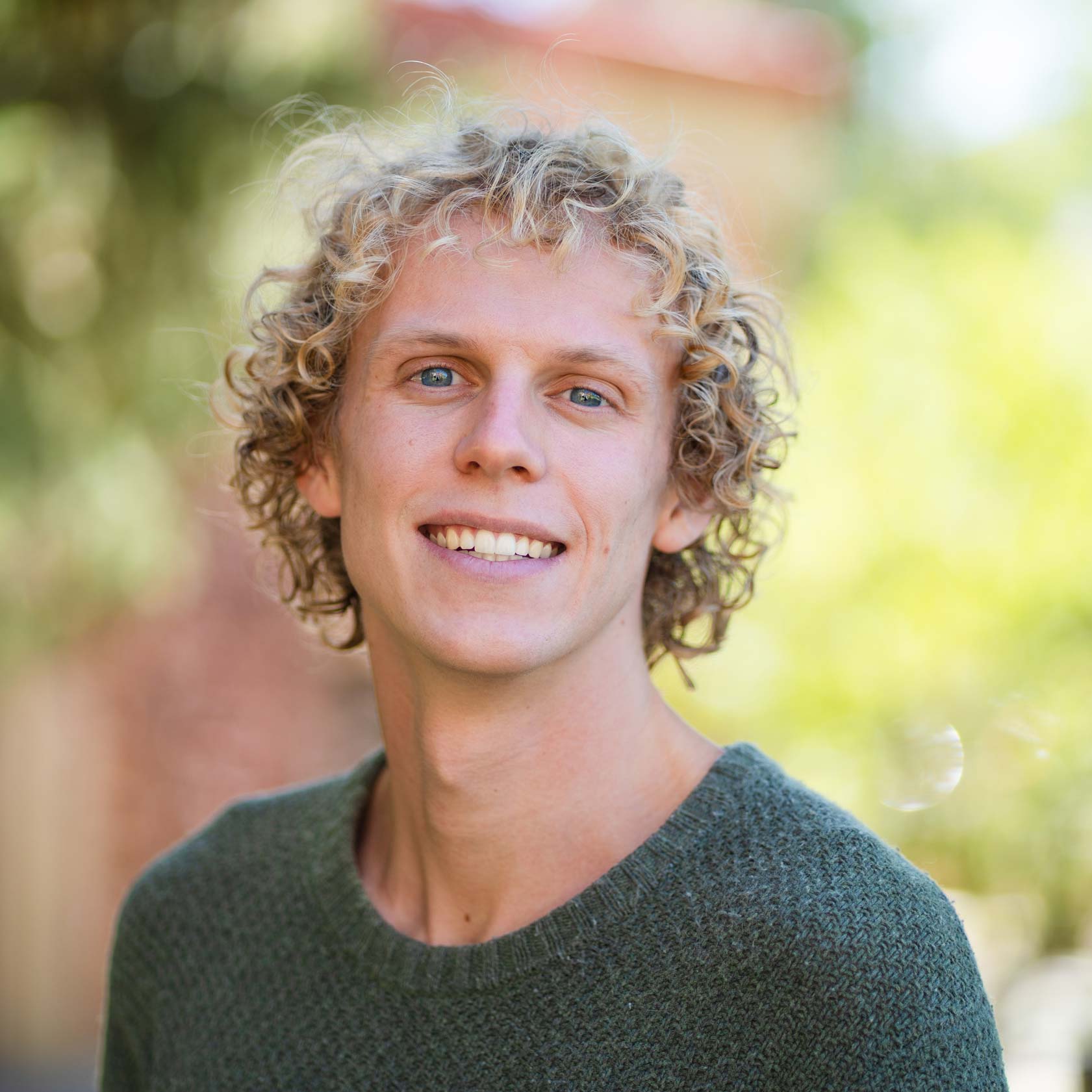 A person with curly blonde hair and blue eyes smiles warmly while wearing a dark green sweater outdoors.