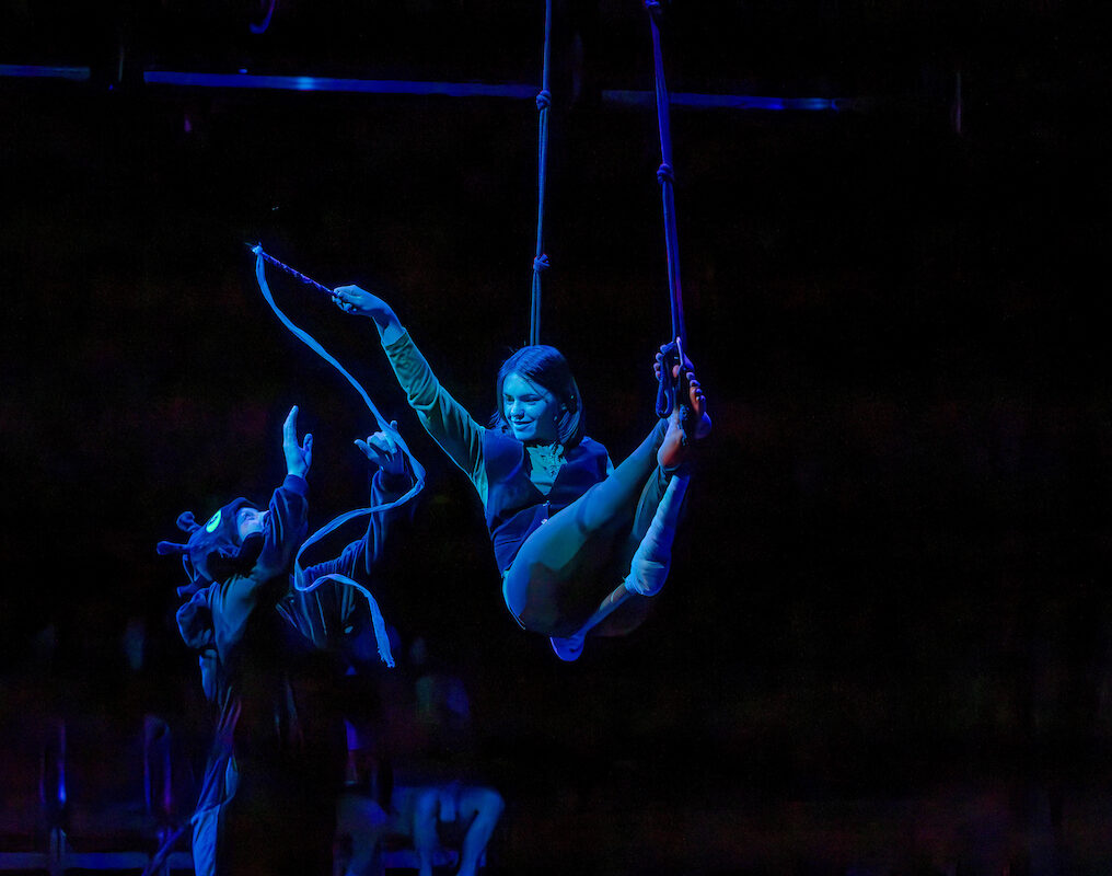 Under blue stage lighting, a smiling aerial performer on a trapeze reaches toward a costumed figure on the ground who is handing them a rope.