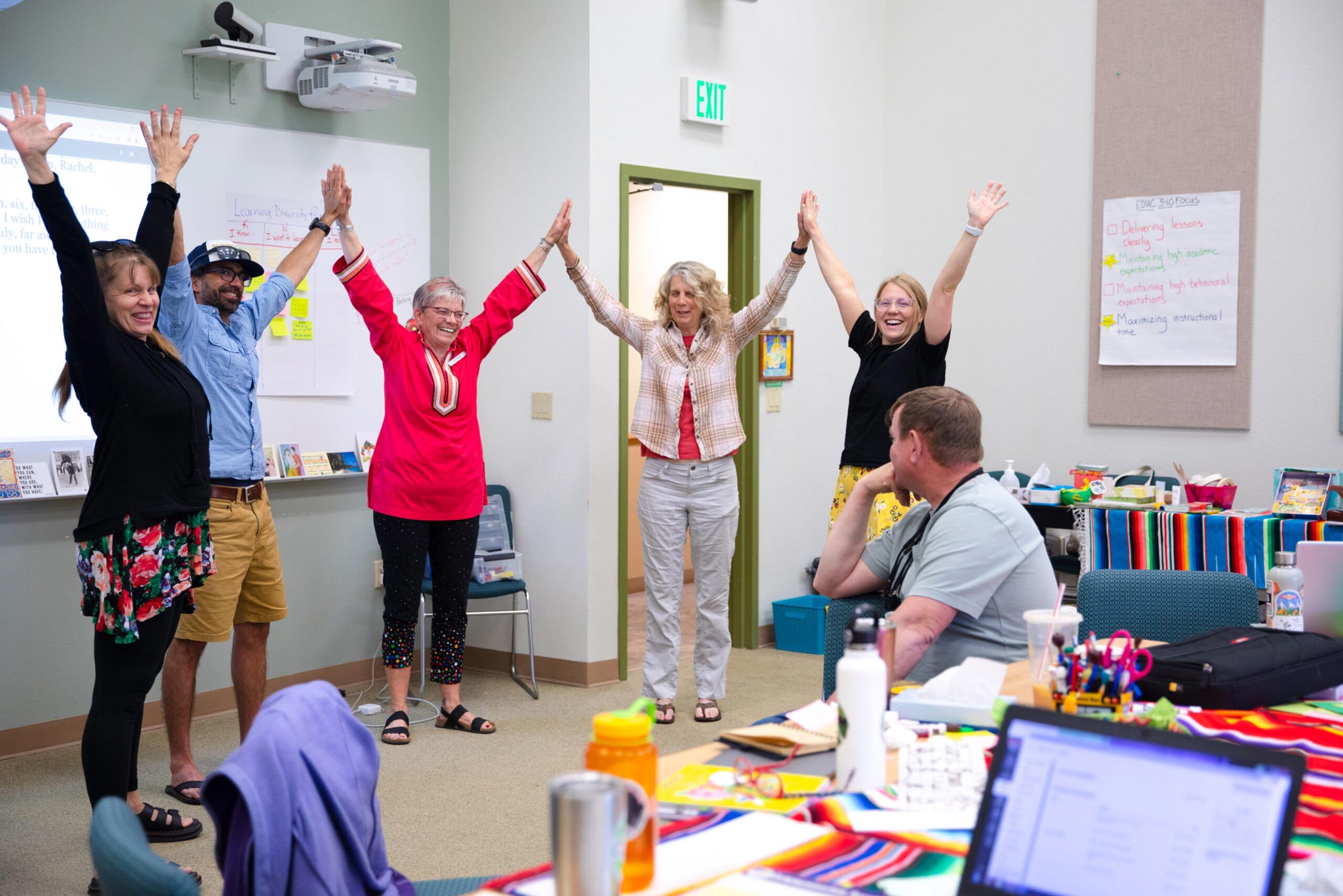 Some students and an instructor stand in a half circle and stretch their arms up toward the ceiling at the front of the classroom.