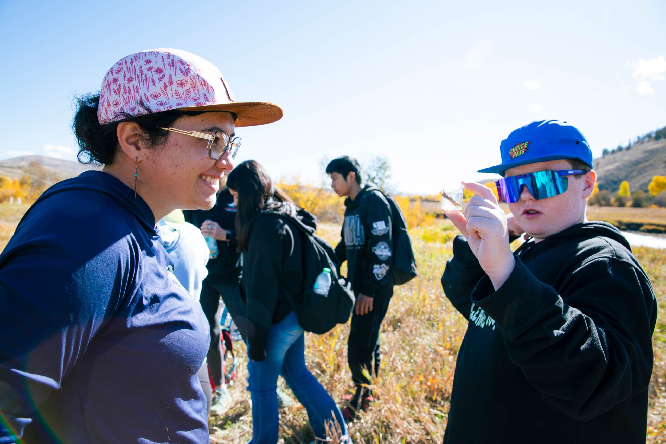 Ecology and Environmental Management graduate student, Sophia Reggiani, smiles as a boy holds up a small plastic vessel containing an aquatic insect in a sample of water.