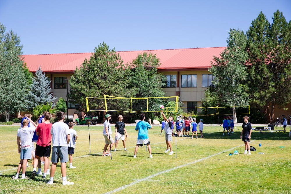 A group of young people play volleyball on a sunny day in a grassy field, with a building and trees in the background.