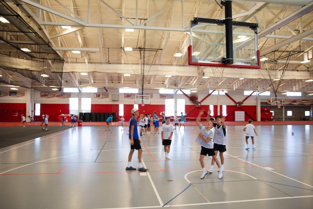 Young people play basketball in a large indoor gymnasium with multiple hoops and red accents.