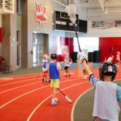 A young athlete in a helmet and jersey raises a lacrosse stick high on an indoor track, with other players practicing nearby.